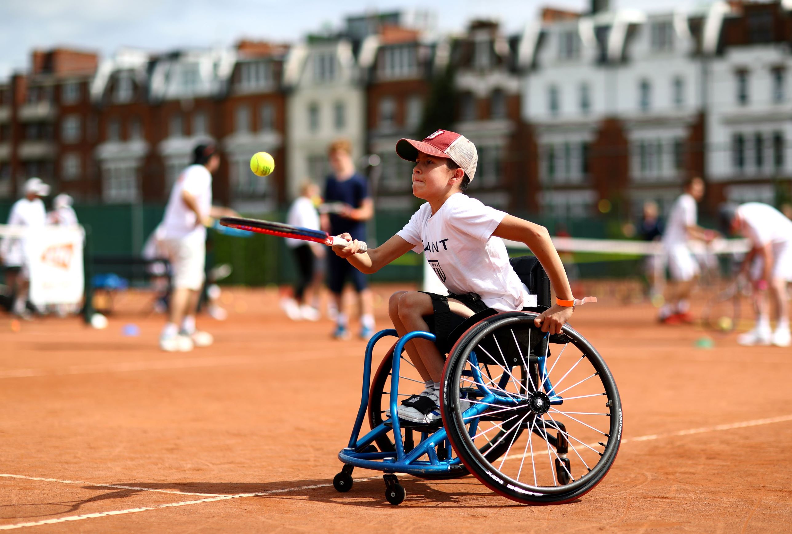 A young boy in a wheelchair doing keepie uppies with a tennis racket and ball on a clay court