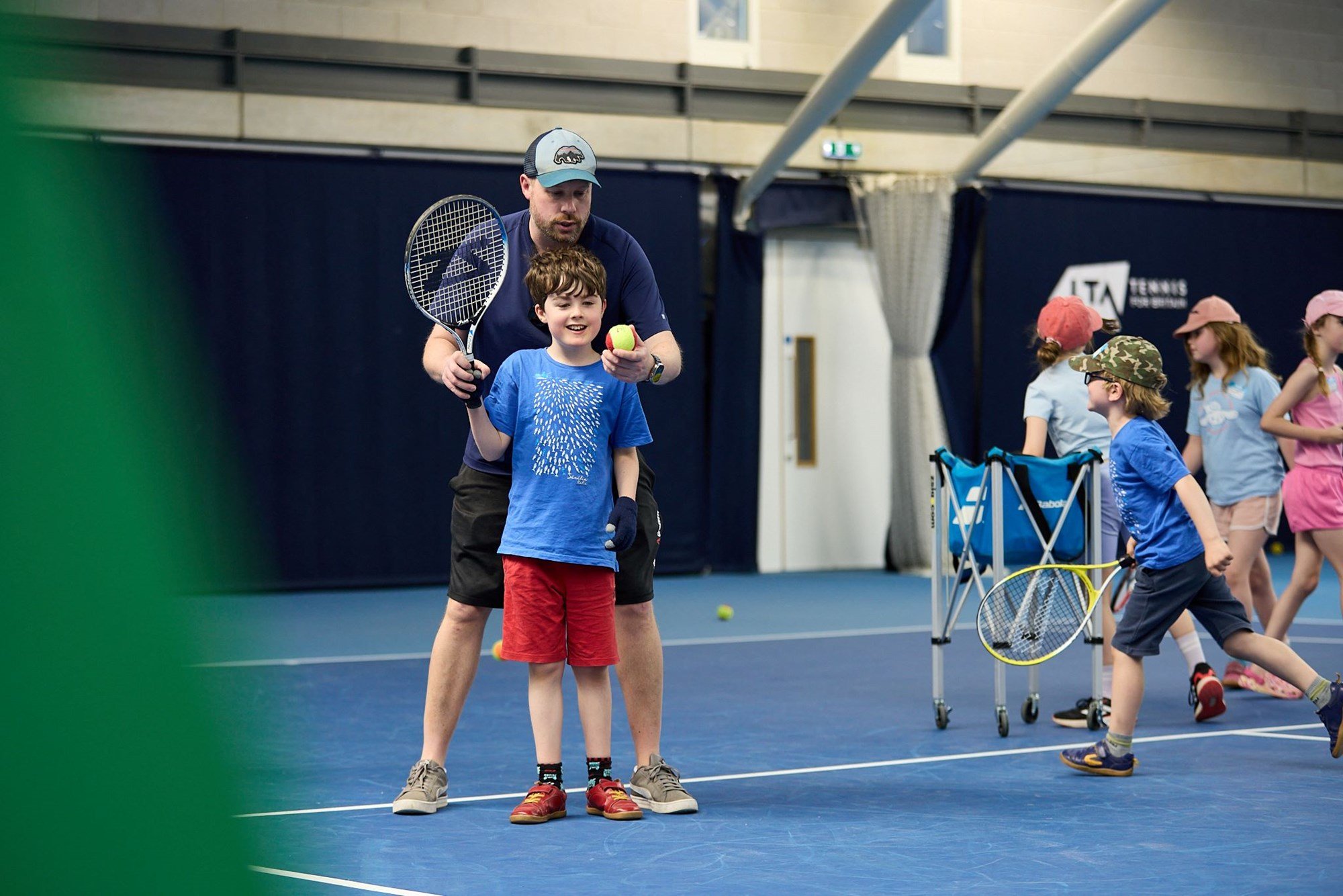 A coach standing behind a young player guiding the racket in the boy's hand while holding up a tennis ball