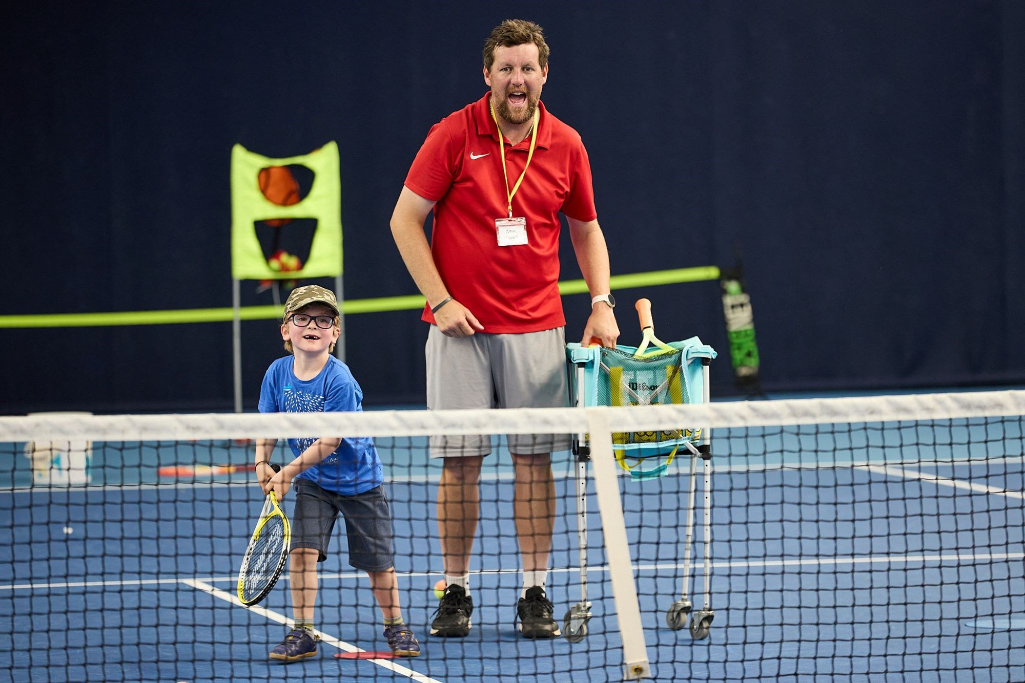 A coach and young male player behind a net, the young boy is hitting balls with his racket