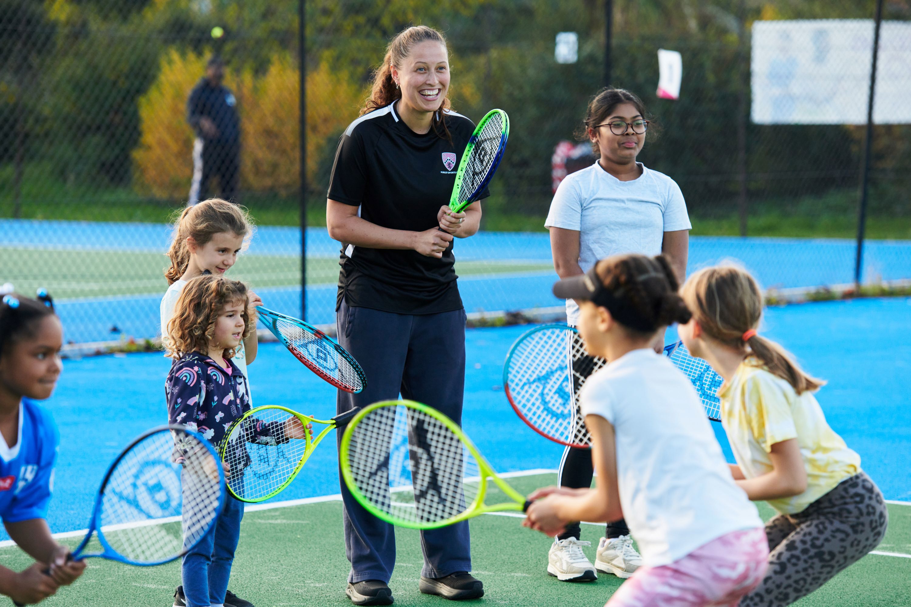 A coach on court, holding a racket and smiling while a group of young players complete a drill with rackets in front of her