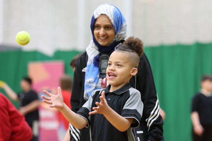 A coach watching on, smiling as a young boy with a hearing aid catches a tennis ball