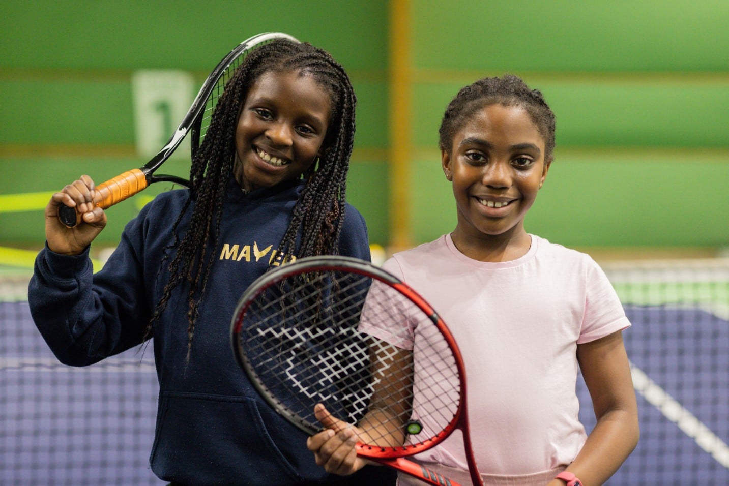 Two girls stand side-by-side smiling and holding tennis rackets, in front of a tennis net