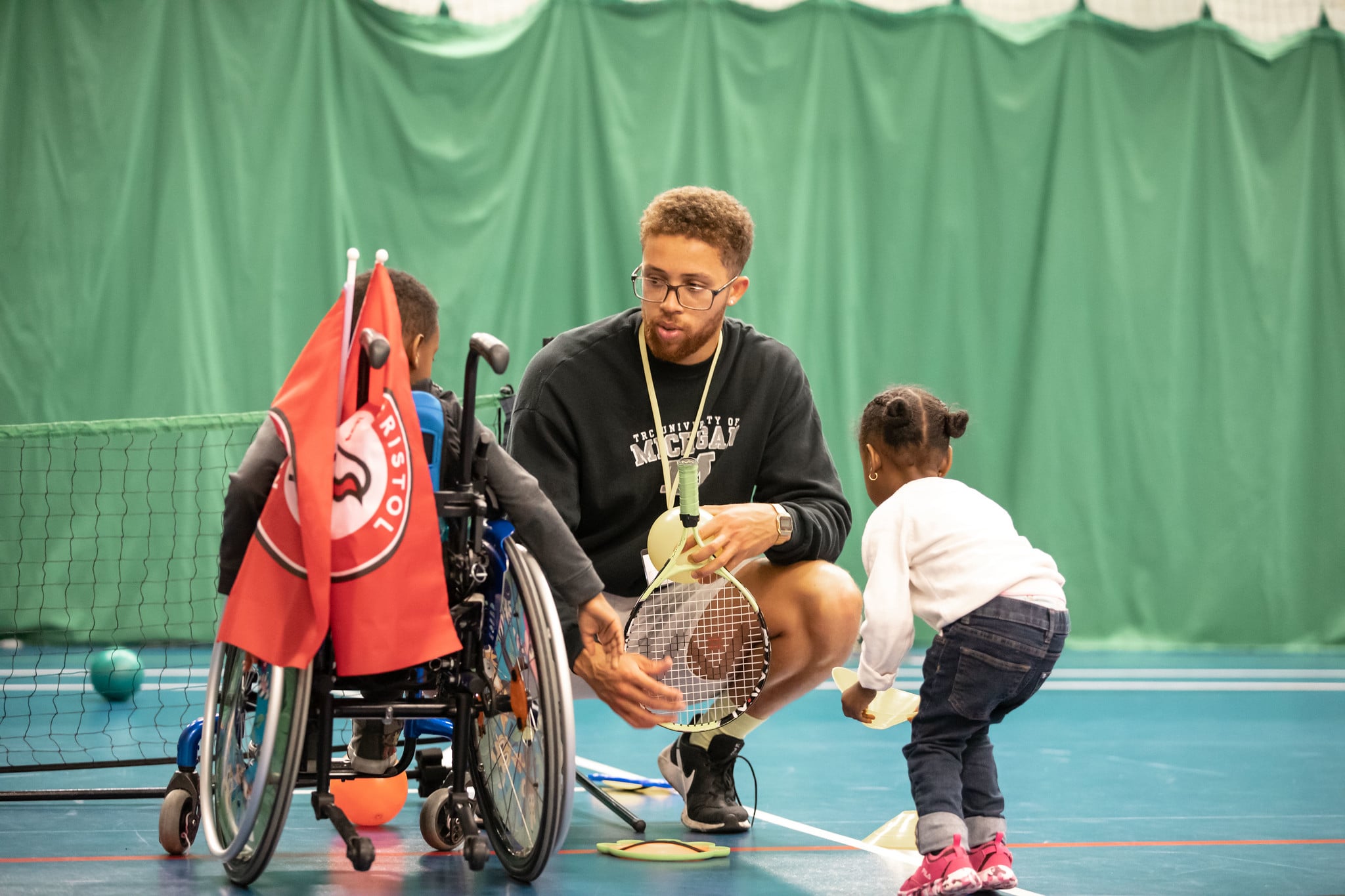 A man crouches to talk to two young children. He is holding a tennis racket, while the child on his right facing him is in a wheelchair, and the child on his left picks up a cone