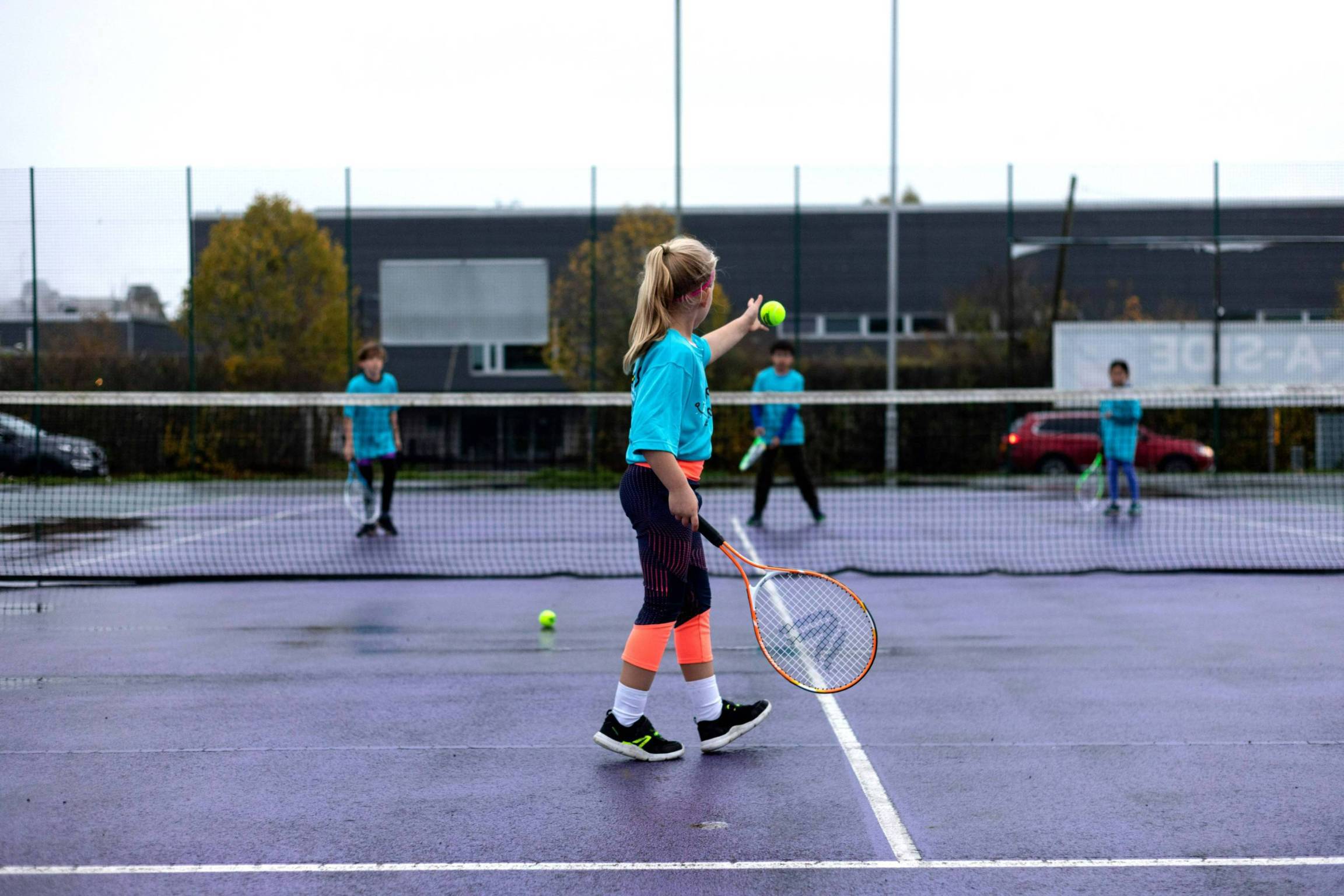 A young girl drops a tennis ball and preapres to hit it over a tennis net in the direction of three other children stood on the other side of the net