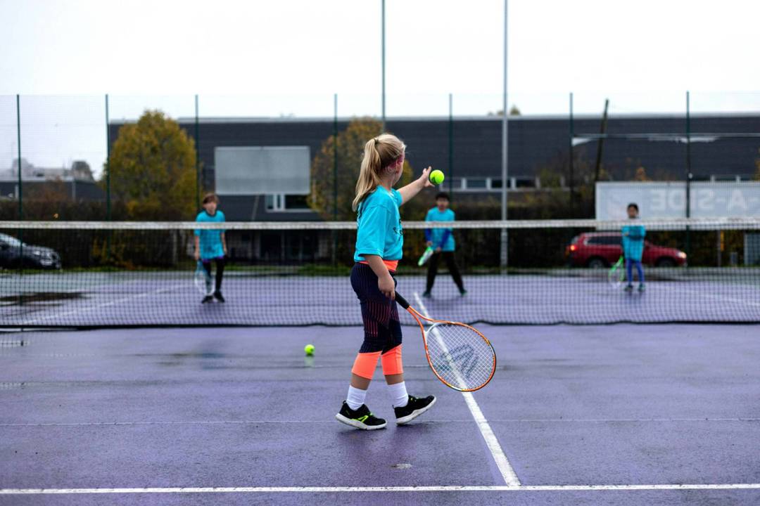 A young girl drops a tennis ball and preapres to hit it over a tennis net in the direction of three other children stood on the other side of the net