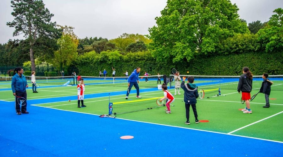 A mixture of children and adults play tennis on tennis courts in a park. The courts are green, with blue surrounds.