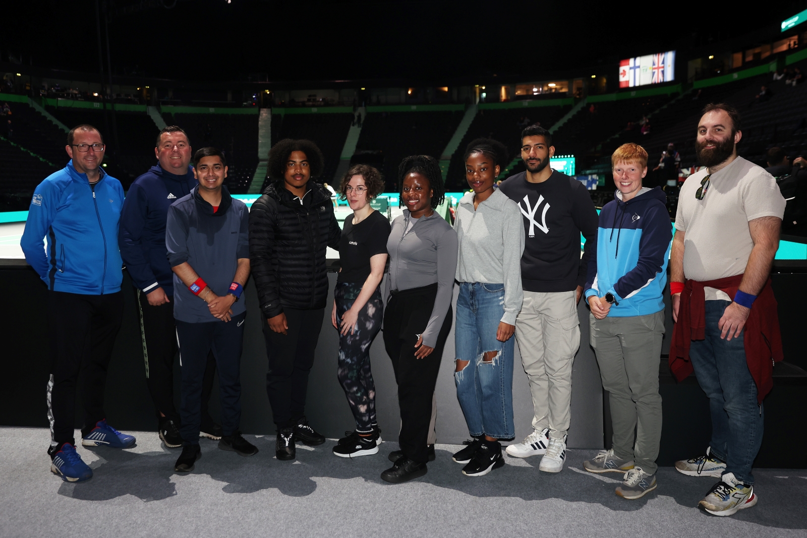 A group of young people stand against a barrier, with a large area of arena seating visible behind them