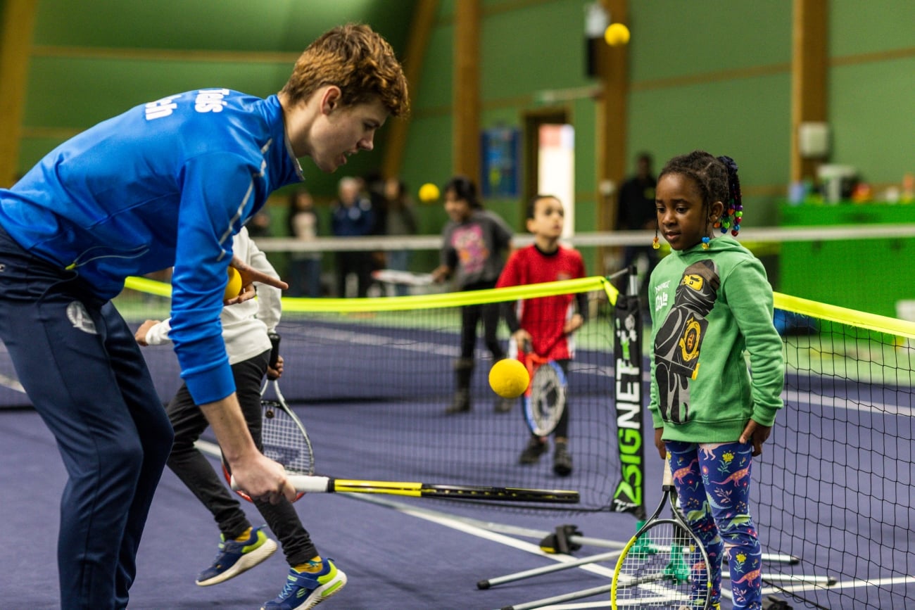 An instructor in blue helps young children play tennis on an indoor court with mini nets and soft balls.