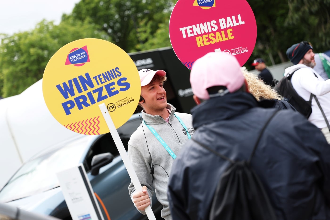 A male staff member holding a tennis ball resale sign and a win tennis prizes sign
