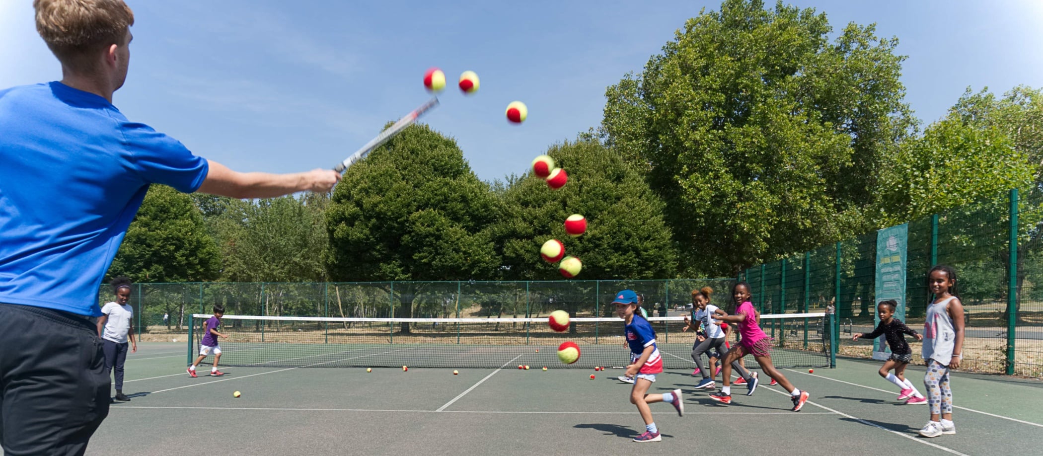 An instructor hitting tennis balls towards a group of children with their rackets