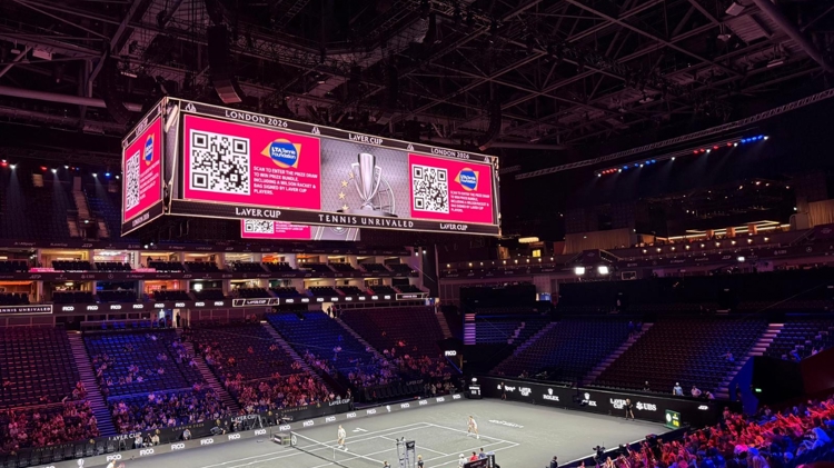 Large digital screen displaying QR code and LTA Tennis Foundation logo, hanging above a tennis court inside an area 