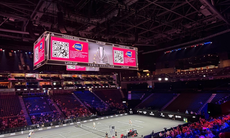 Large digital screen displaying QR code and LTA Tennis Foundation logo, hanging above a tennis court inside an area 