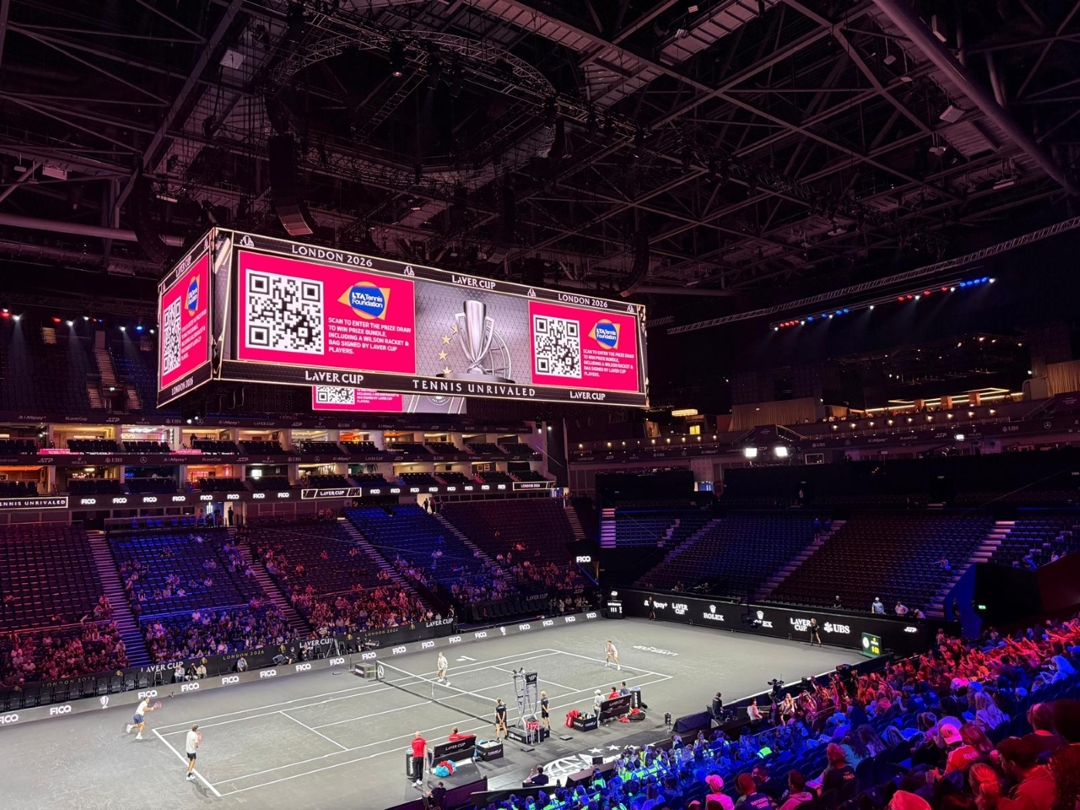 Large digital screen displaying QR code and LTA Tennis Foundation logo, hanging above a tennis court inside an area 
