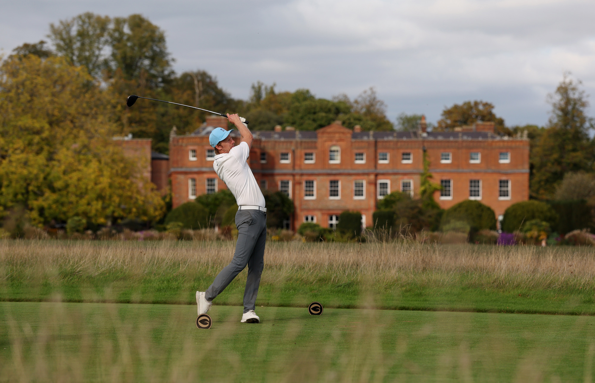 A man poses after hitting a golf ball with a large house in the background
