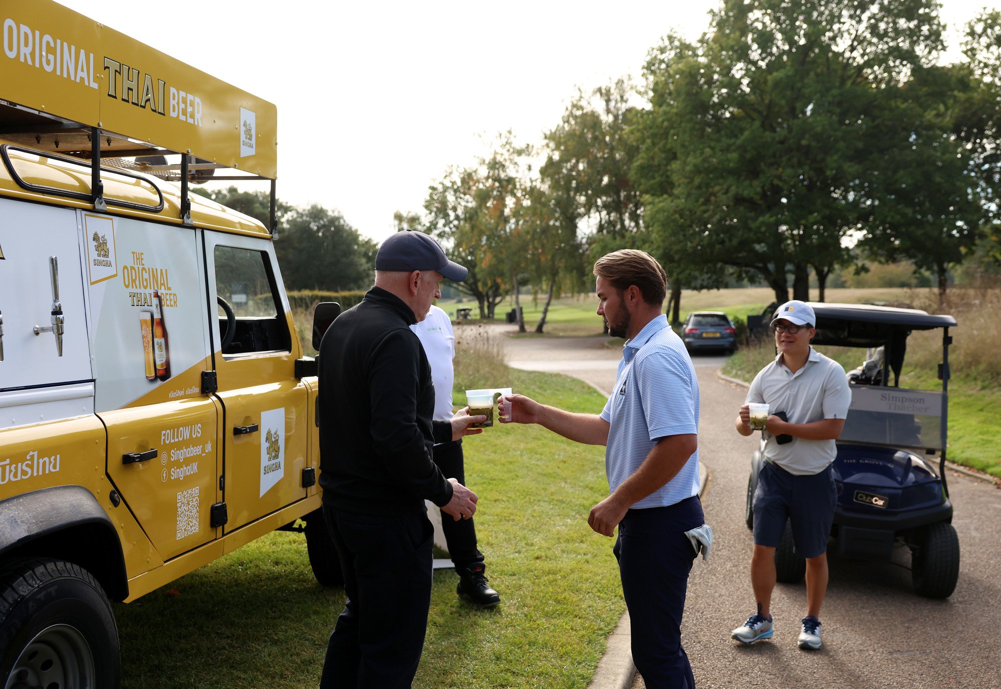 A man hands another man a beer in front of a Singha Van