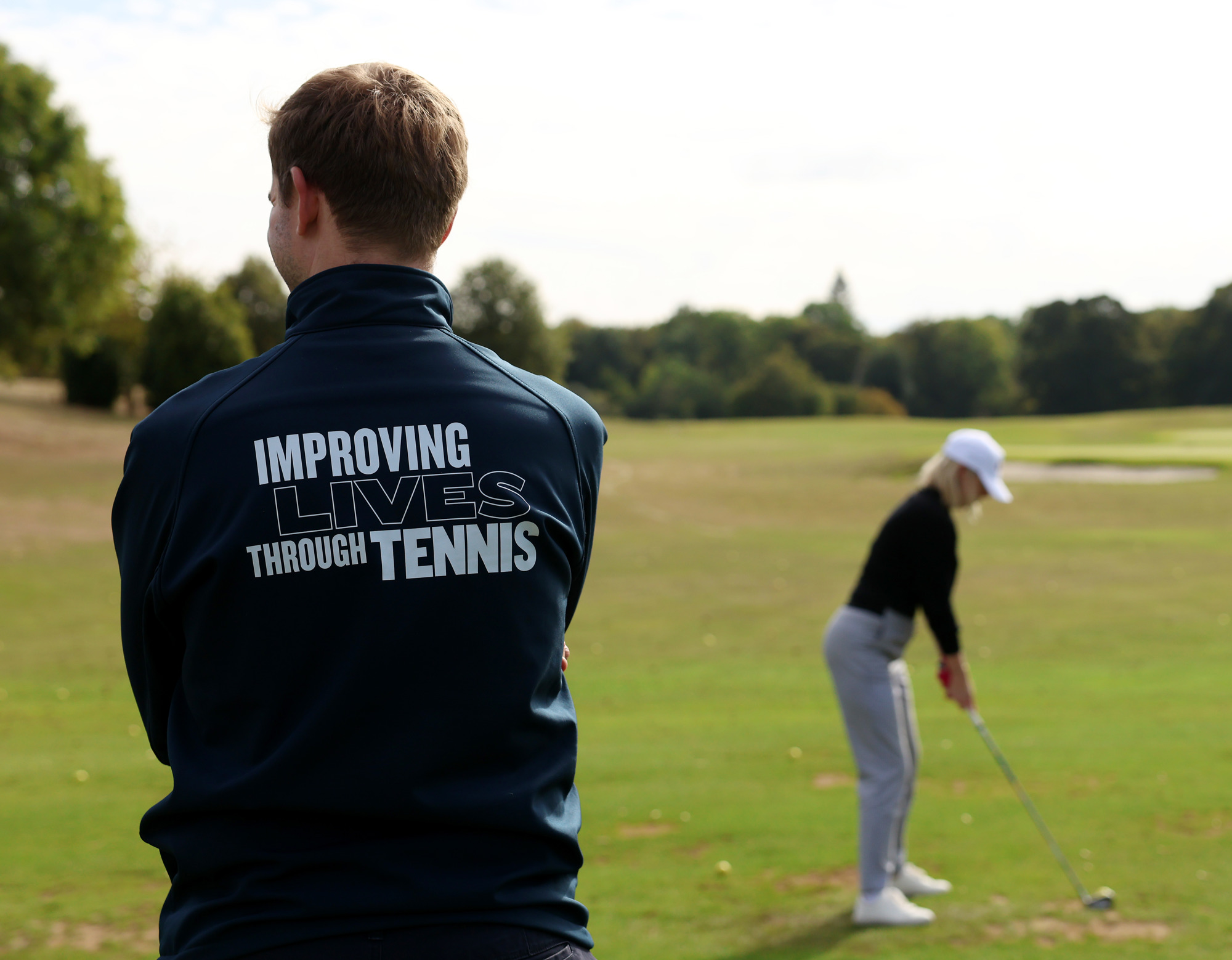 A golfer swings in the background with a volunteer stood in the foreground