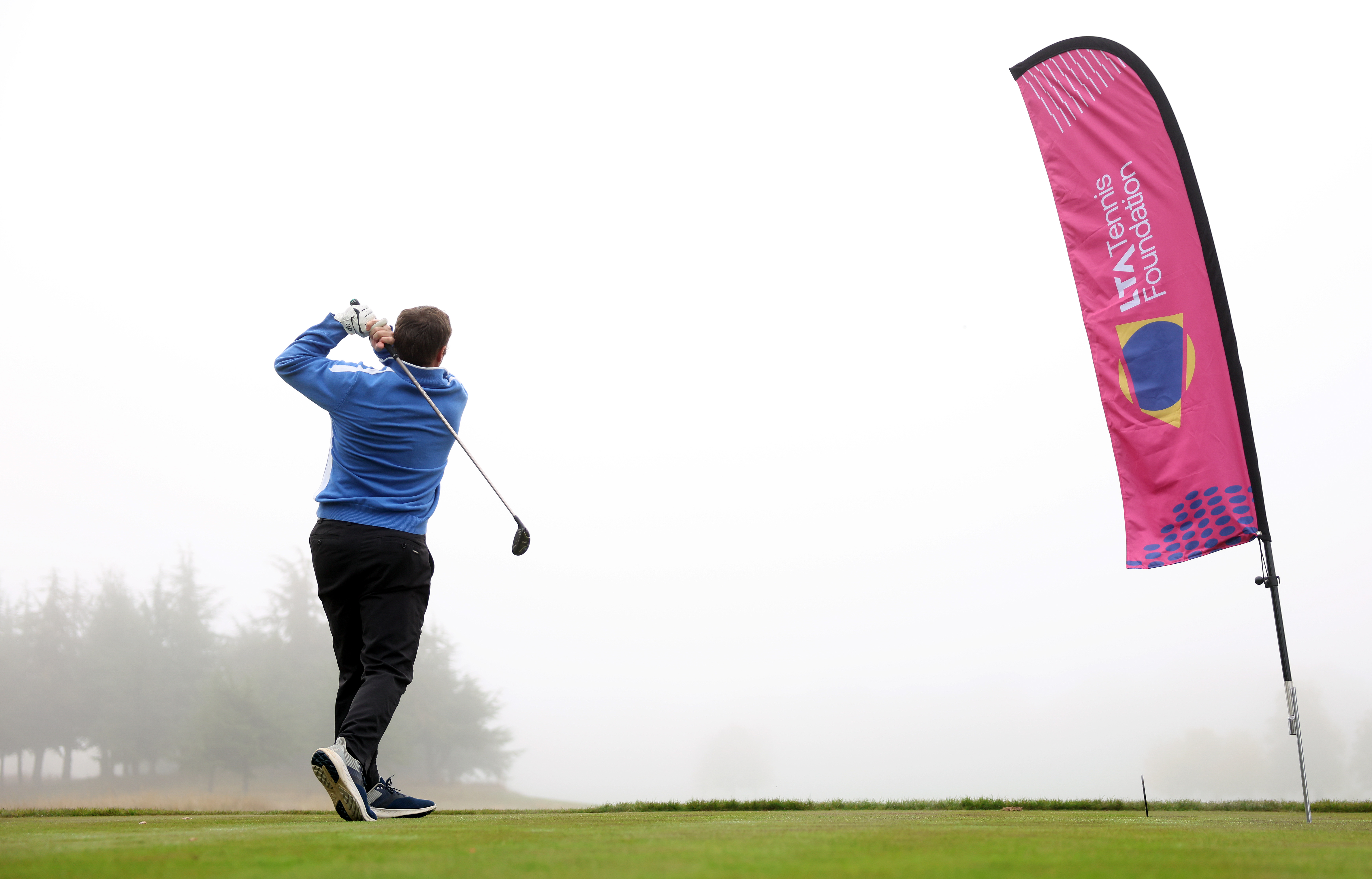 A man poses after hitting a golf ball with an LTA Tennis Foundation feather banner next to him