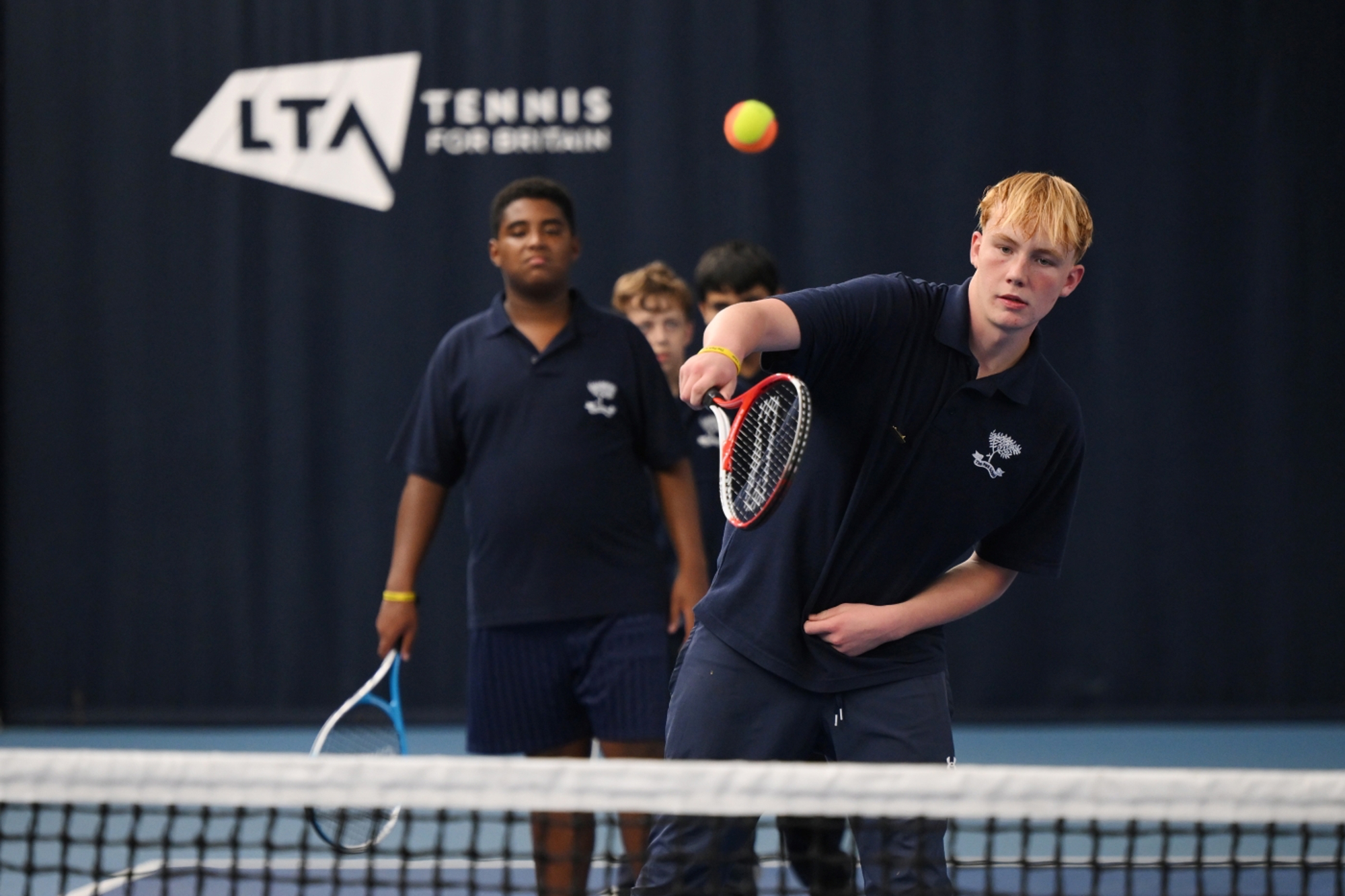 A young person with a tennis racket at the net on a tennis court hitting the ball. Another young person stands behind waiting to have a turn