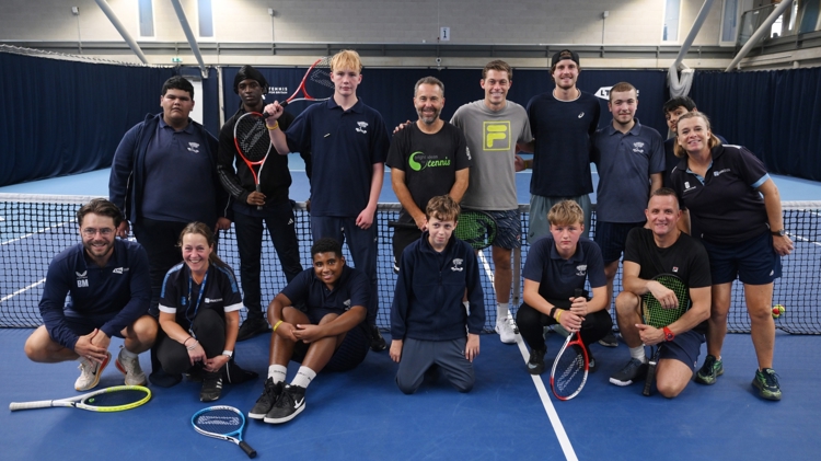 A group of 9 young people with two coaches stood in front of the net on a tennis court with professional tennis players Neil Skupski and Tobey Samuel