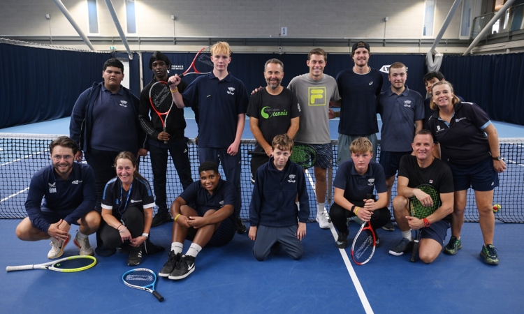 A group of 9 young people with two coaches stood in front of the net on a tennis court with professional tennis players Neil Skupski and Tobey Samuel