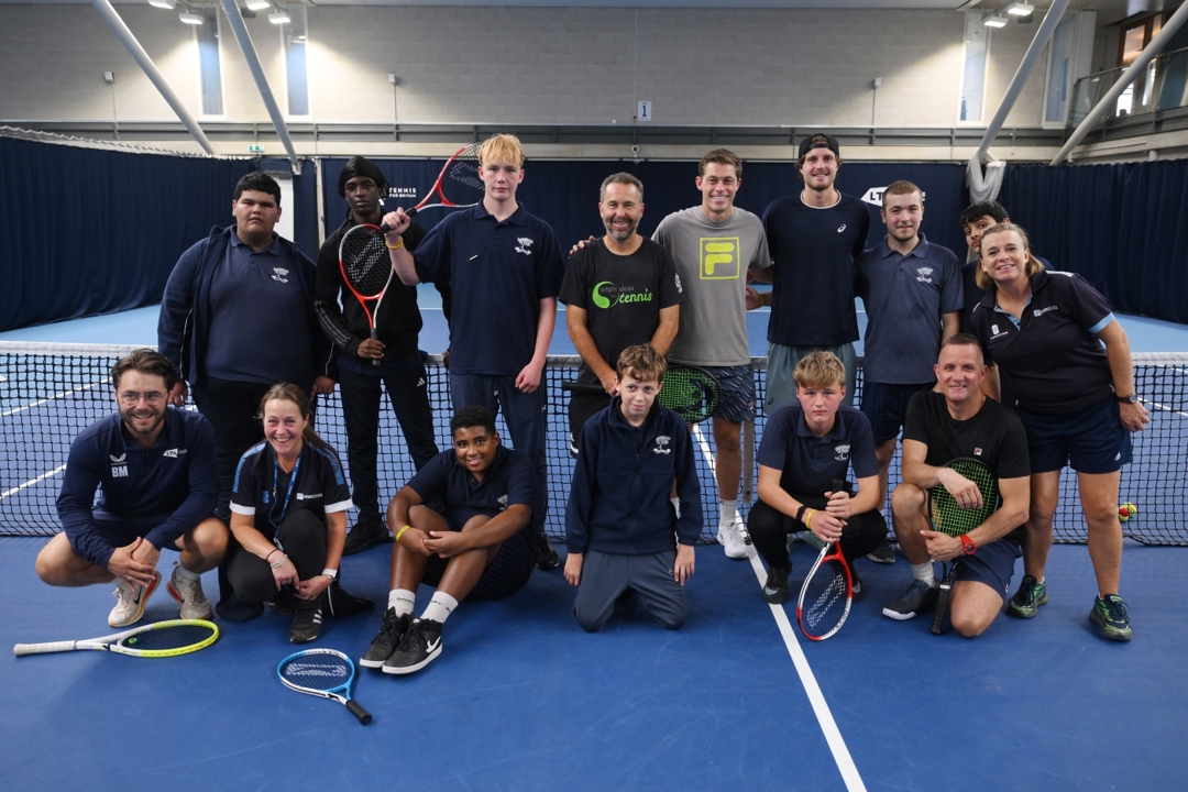 A group of 9 young people with two coaches stood in front of the net on a tennis court with professional tennis players Neil Skupski and Tobey Samuel