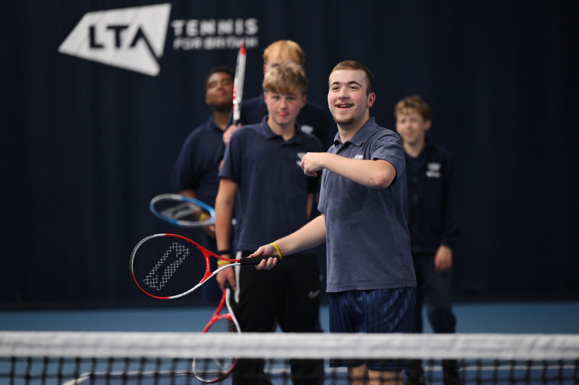 A young player stood at the net on a tennis court waiting to hit a ball, smiling at the camera