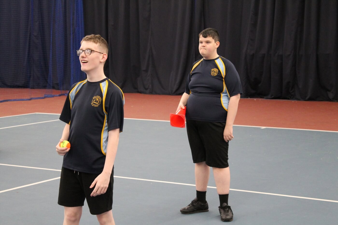 Two students on a tennis court at an indoor centre. One is holding a ball and one is holding a cone ready to play a tennis skills game