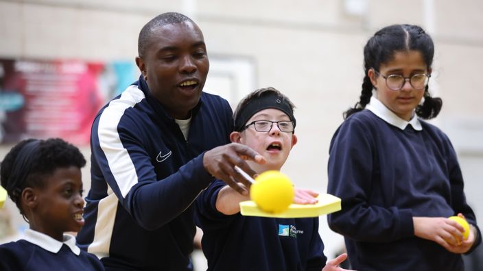 A coach standing behind a young boy, helping him hit a soft ball with a padel